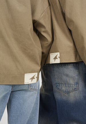 Deux personnes portant des vestes beige et des jeans bleus, montrant les étiquettes des vestes avec un logo en forme d'étoile près de l'ourlet inférieur.