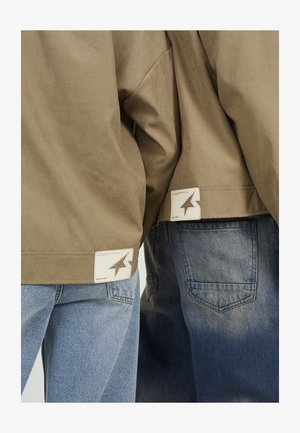 Deux personnes portant des vestes beige et des jeans bleus, montrant les étiquettes des vestes avec un logo en forme d'étoile près de l'ourlet inférieur.
