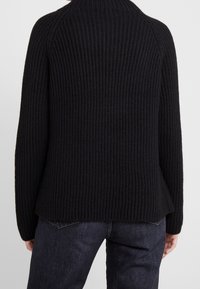 Suéter negro acanalado con cuello alto y mangas largas, con un ajuste holgado y ligero vuelo en el dobladillo. Textura suave.