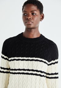 Maglione lavorato a maglia a trecce nero e crema con scollatura rotonda. Presenta strisce orizzontali alternate di filato nero solido e crema.