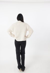 Suéter texturizado de color crema con un cuello alto, combinado con jeans negros ajustados. La modelo está de pie con las manos en las caderas frente a un fondo simple.