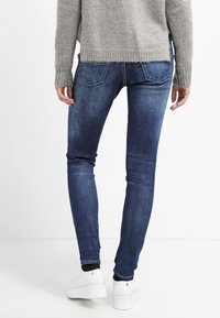 Person som bär urblekta blå skinny jeans, beige stickad tröja och vita plattformssneakers, sedd bakifrån mot en vit bakgrund.