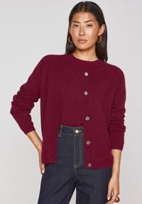 Cardigan bordeaux con collo a giro, chiusura con bottoni, polsi a costine e texture lavorata a maglia, abbinato a jeans alto in vita di denim scuro.