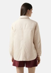 Beżowa pikowana kurtka z ostro zakończonym kołnierzem, długimi rękawami i detalem w postaci szwu z tyłu. Połączona z bordowymi szortami, posiada mankiety zapinane na guziki.