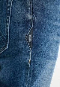 Jeans in denim blu scuro con un leggero sbiadimento. Presentano un distintivo patch con logo sulla tasca frontale e dettagli di cucitura ben visibili.