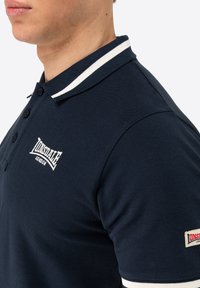 Marineblå polo-shirt med en tekstureret finish, hvidstribede krave, tre-knaps placket og broderet logo på brystet og ærmet.