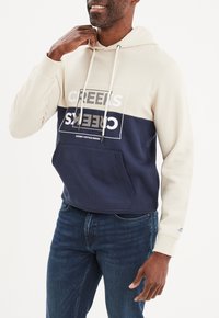 Sudadera con capucha de sección superior beige y sección inferior navy, que cuenta con un bolsillo frontal y un estampado gráfico blanco. Fabricada con tela suave y con puños acanalados.