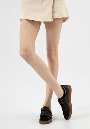 Nohy zkřížené, oblečené v béžových šortkách a černých teniskách s třemi suchými zipy a hnědými podrážkami na bílém pozadí.
