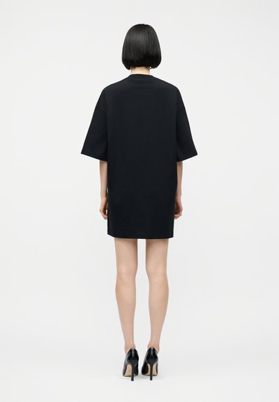 Γυναίκα με κοντά μαύρα μαλλιά, που φοράει χαλαρό μαύρο φόρεμα και μαύρα παπούτσια με ψηλό τακούνι, στέκεται με την πλάτη γυρισμένη σε απλό φόντο.