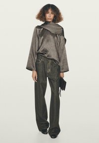 Žena s kudrnatými vlasy nosí volný metalicky šedý top, široké tmavé kalhoty, černé sandály na otevřenou špičku a v ruce drží malou černou kabelku.