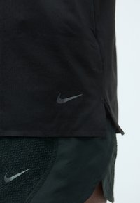 T-shirt de sport Nike noir à manches courtes, avec une texture subtile et un logo gris sur le côté gauche. Assorti avec un short vert.