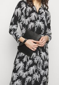 Černá kožená kabelka s texturovaným povrchem, se zápěstním popruhem. Nositá s černými šaty zdobenými bílými vzory palmových listů.