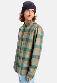 Camisa de franela a cuadros en verde y marrón, con mangas largas, cierre de botones y bolsillo. Combinada con un gorro de punto azul.