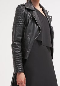 Czarna skórzana kurtka motocyklowa z pikowanymi akcentami na ramionach, zapinana na zamek, o skróconym fasonie, noszona na czarnej sukience. Gładka tekstura.