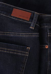 Sötét farmernadrág bőr címkével, amelyen a "TATUUM" felirat olvasható. Narancssárga varrással, egy hátsó zsebbel és réz színű fém kiegészítőkkel rendelkezik.