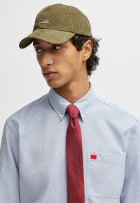 Olivengrøn fløjlskappe med "HUGO" broderet på forsiden, båret med en lyseblå skjorte med krave og et rødt mønstret slips.