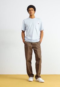 Lyseblå bomull T-skjorte med lomme, kombinert med beige bukser. Modellen har på seg hvite sneakers og står mot en ensfarget bakgrunn.