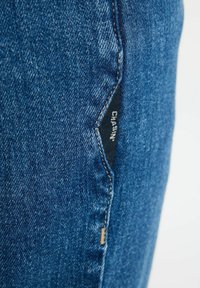 Sinised denim püksid, millel on lähedal vaatamine mustast etiketist, millel on külgõmblusel kirjas "CHASIN'", näha on õmbluseid ja tekstureeritud kangast.