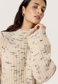Strickpullover in Creme mit schwarzen Sprenkeln, geripptem Muster, hohem Kragen und weiten Ärmeln. Hergestellt aus weicher Wolle für zusätzlichen Komfort.