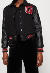 Svart varsityjacka med ullkropp och lämskinn. Har en röd bokstavs"G"-applikation och randiga ribbade muddar. Utrustad med tryckknappar.