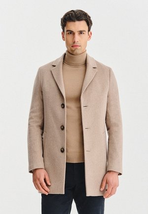 Beżowy wełniany płaszcz z kołnierzem, z trzema czarnymi guzikami, z jednym rozcięciem z tyłu i teksturowanym wykończeniem. Nałożony na beżowy golf.