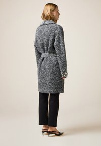 Grau strukturierter Mantel mit tailliertem Gürtel und Schalkragen, kombiniert mit schwarzen Slim-Hosen und schwarzen Absatzschuhen. Rückansicht.