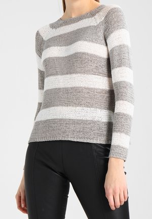 Femme portant un pull en maille à manches longues avec des rayures horizontales grises et blanches et un pantalon noir ajusté sur un fond uni.