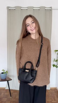 Črna usnjena torbica z dvema ročajema in snemljivim paščkom, z teksturirano površino in zlato obarvanimi kovinskimi deli, kombinirana z rjavim puloverjem.