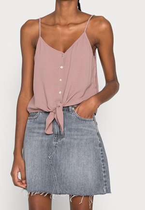 Donna che indossa un top senza maniche rosa polvere con bottoni e laccio frontale e una gonna di jeans grigia sbiadita con orlo sfrangiato.