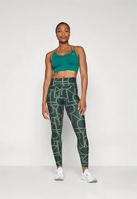 Sportinė liemenėlė žalsvai mėlynos spalvos su plonais dirželiais, derinama su žaliomis tampris su abstrakčiu geometriniu raštu. Baltos sportbačiai užbaigia aprangą.