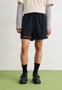 Person som bär svarta Nike-shorts med orange tryck, svarta strumpor, svarta spänneskor och en beige tröja med grå randiga långärmar.