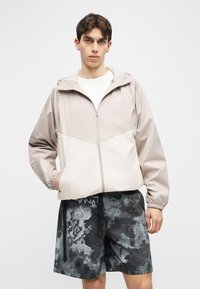 Νέος άνδρας που φοράει μπεζ και λευκό φερμουάρ με κουκούλα, και μαύρα-γκρι σορτς με μοτίβο, στέκεται με το ένα χέρι στην τσέπη του.