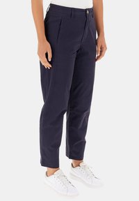 Pantaloni navy con un design sartorial, caratterizzati da pieghe frontali, chiusura con bottone e orli risvoltati. Abbinati a scarpe da ginnastica bianche.