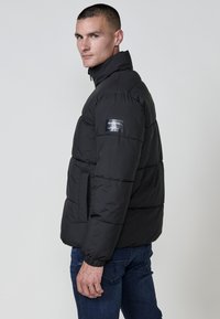 Sort pufferjakke med høj krave, vandret syning og et mærke på venstre ærme. Båret over blå denim jeans.