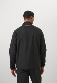 Svart jacka med hög krage och elastiska ärmslut; Reebok-logotyp i rött på övre ryggen; slitstarkt och lätt material.