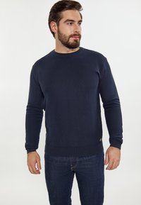 Sötétkék kötött pulóver, kerek nyakkal, bordás mandzsettákkal és aljjal. Sötétkék farmernadrággal viselve, amelynek az alján egy kis logó részlet található.