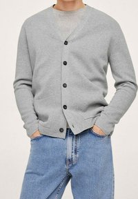 Cardigan gris en matière douce, avec un col en V, six boutons noirs, deux poches devant et une coupe confortable portée sur un T-shirt gris.