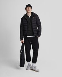 Zwarte gewatteerde jas met capuchon, groene sweatshirt, zwarte joggers, grijze sneakers en een zwarte tote bag. Model staat tegen een grijze achtergrond.