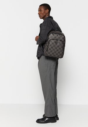 Zaino nero con motivo geometrico a triangoli, dotato di scomparti con cerniera e tessuto resistente, indossato da una persona con un abbigliamento grigio.