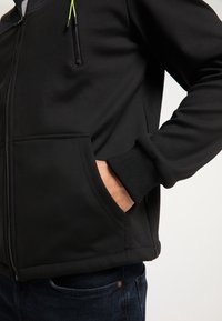 Osoba w czarnej kurtce zapinanej na suwak, z ręką w przedniej kieszeni, w ciemnoniebieskich dżinsach, pokazana od w talii do połowy ramienia.