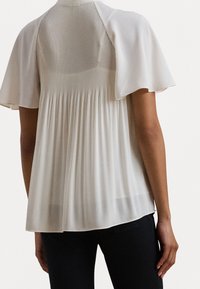 Blouse blanche avec dos plissé, manches évasées et texture lisse. Tissu doux avec un éclat subtil, idéal pour être superposé à d'autres vêtements.
