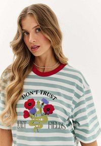 Pruhované tričko v mátově zelené a bílé barvě, s červeným límcem. Hlavní grafika obsahuje květiny a hada s textem: "NEVĚŘTE TICHO POLÍ."