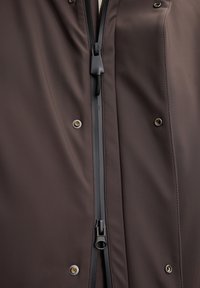 Nahaufnahme der Vorderseite einer braunen Jacke mit einem schwarzen Zwei-Wege-Reißverschluss und vier metallischen Druckknöpfen auf wasserdichtem Stoff.