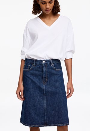 Femme portant une chemise blanche à manches longues avec col en V, rentrée dans une jupe en jean bleue longueur genoux avec poches avant et fermeture à boutons.