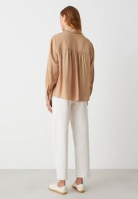 Blusa a maniche lunghe di un leggero beige con una vestibilità rilassata, caratterizzata da pieghe sul retro, indossata con pantaloni bianchi e sneakers beige.