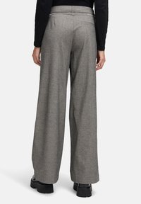 Vrouw in grijze high-waist, wijde broek en zwarte platform schoenen, staand tegen een effen lichte achtergrond.
