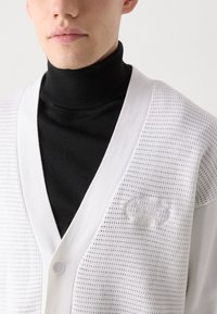 Cárdigan blanco de punto con un escote en V pronunciado, patrón texturizado y un sutil detalle del logo. Llevado sobre un cuello alto negro.