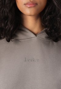 Szürke kapucnis pulóver, puha tapintással, elülső zsebbel és hímezett "LeGer" logóval egy világosabb szürke árnyalatban.