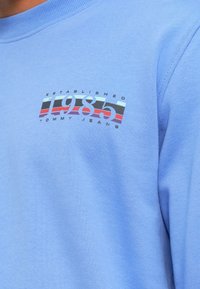 Ανοικτό μπλε φούτερ με γραφικό λογότυπο που απεικονίζει το "1985" και "TOMMY JEANS" σε έντονες, πολύχρωμες ρίγες. Μαλακή υφή, σχέδιο με στρογγυλή λαιμόκοψη.