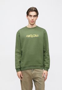 Νεαρός άνδρας με κοντά σκούρα μαλλιά φοράει πράσινο φούτερ με το λογότυπο "Santa Cruz Skateboards" και ανοιχτό καφέ παντελόνι, στέκεται μπροστά σε απλό λευκό φόντο.